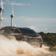 an action photo of a 2024 porsche macan electric prototype. we see the rear of the vehicle as it power-slides across a dirt road, kicking up dust. the vehicle is painted black and wearing vinyl wrapping to disguise its design.